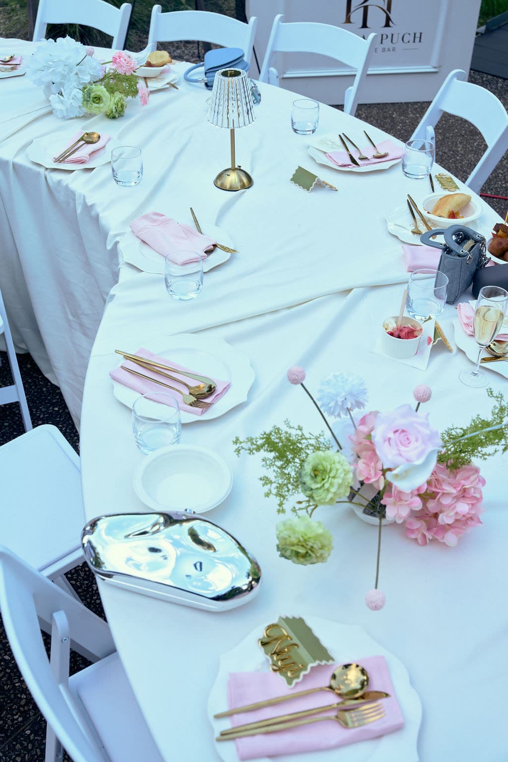 Long outdoor table in white linen with pink napkins, gold cutlery, scalloped plates, a gold cordless lamp, a pink and green hydrangea centrepiece, and gold name cards