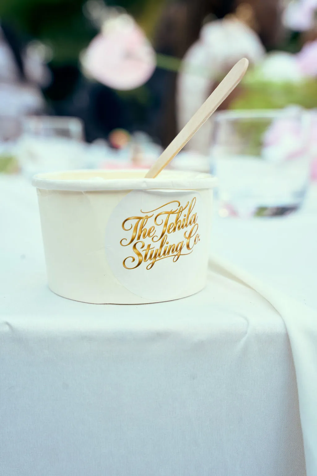 A hand holding a cup of chocolate ice cream with a Tehila Styling Co sticker, beside pink and brown macarons.