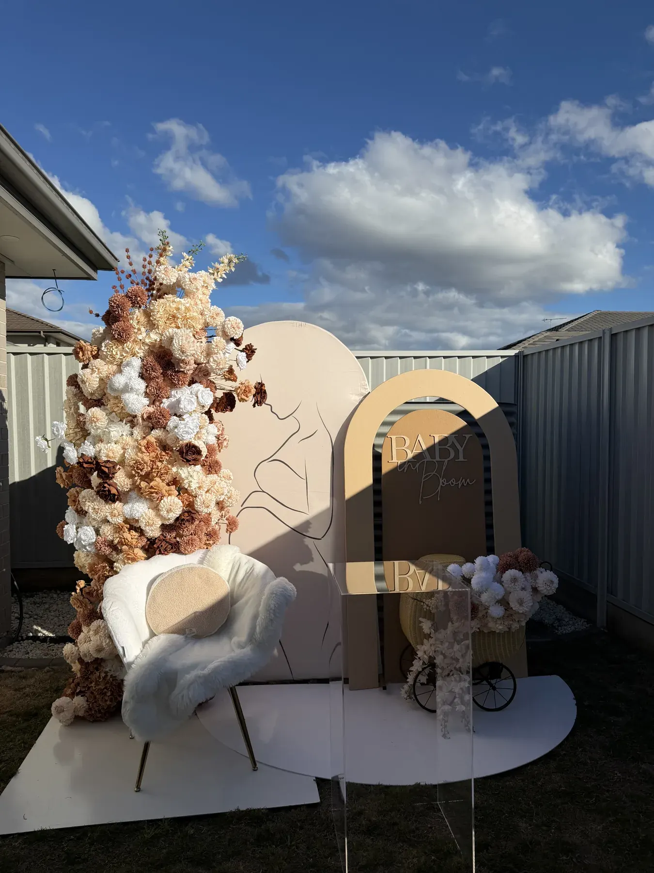 Baby in Bloom arch beside a cream, tan, and brown floral column, a line drawing of a parent and child shown in full, and a white fur chair.