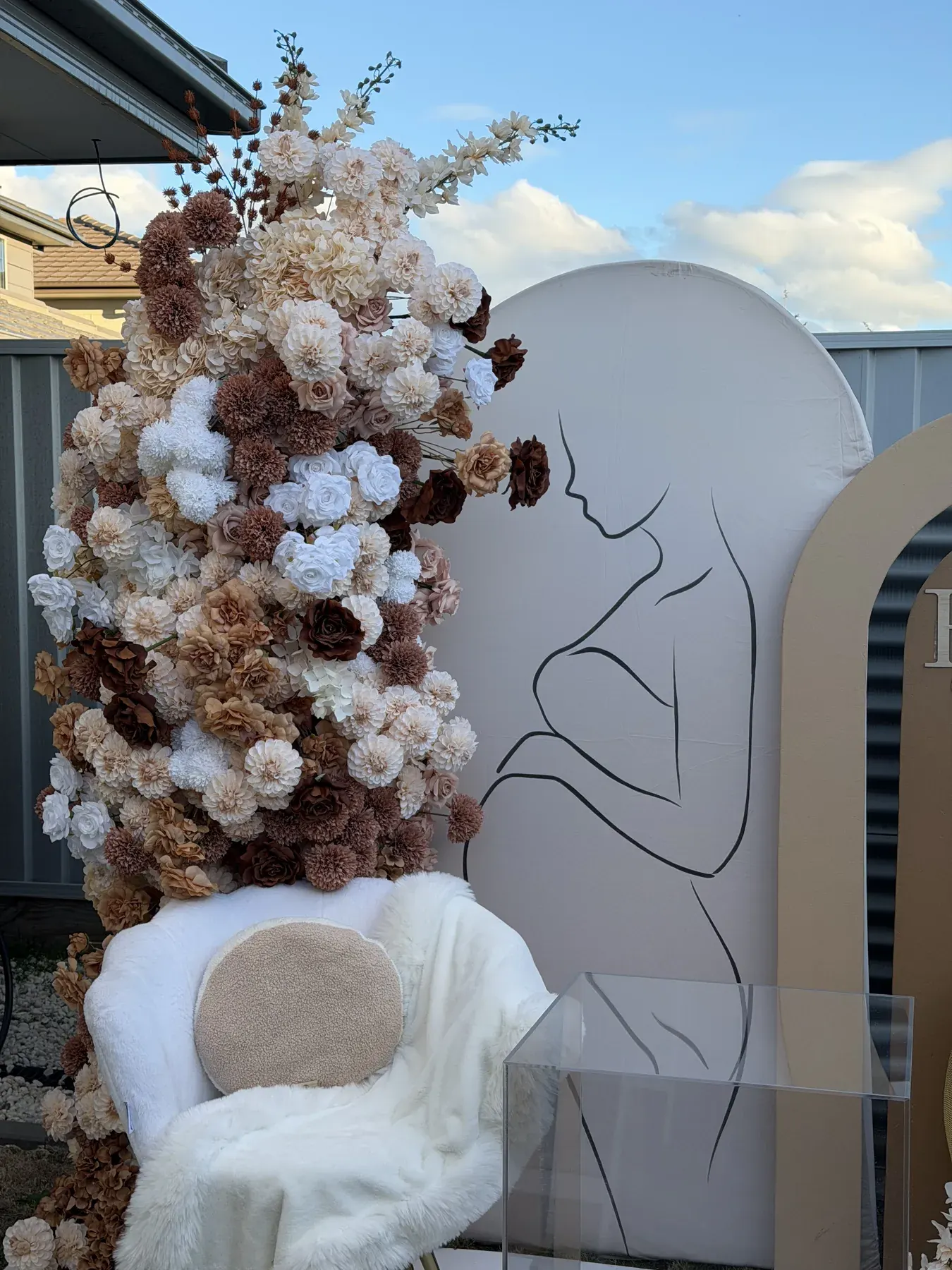 Cream, tan, and brown floral column beside a line drawing of a seated figure, with a white fur chair and a clear plinth.