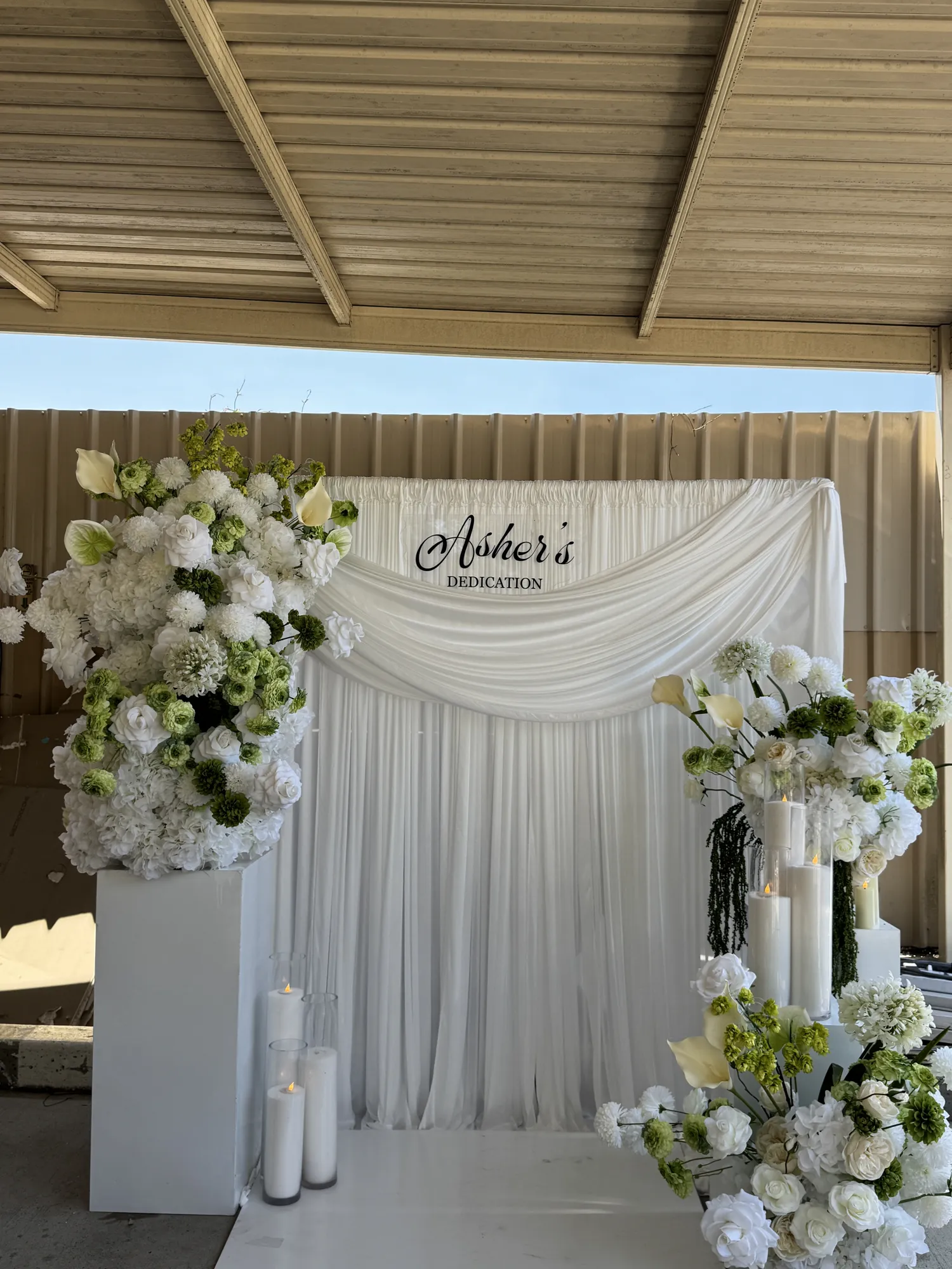A close view of a dedication sign among white flowers, with lit candles in glass holders.