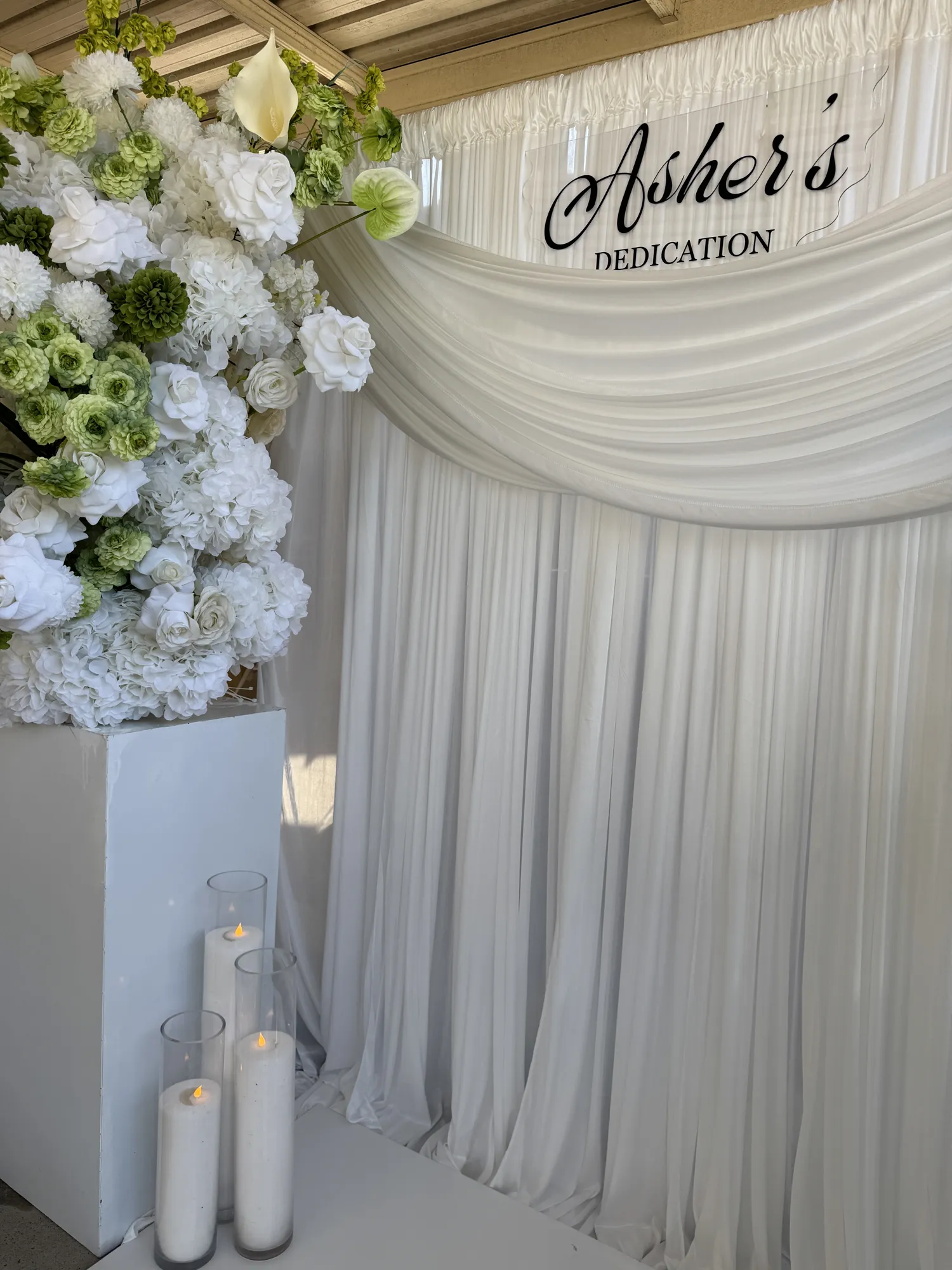 Two guests holding a baby in front of a white and green floral backdrop.