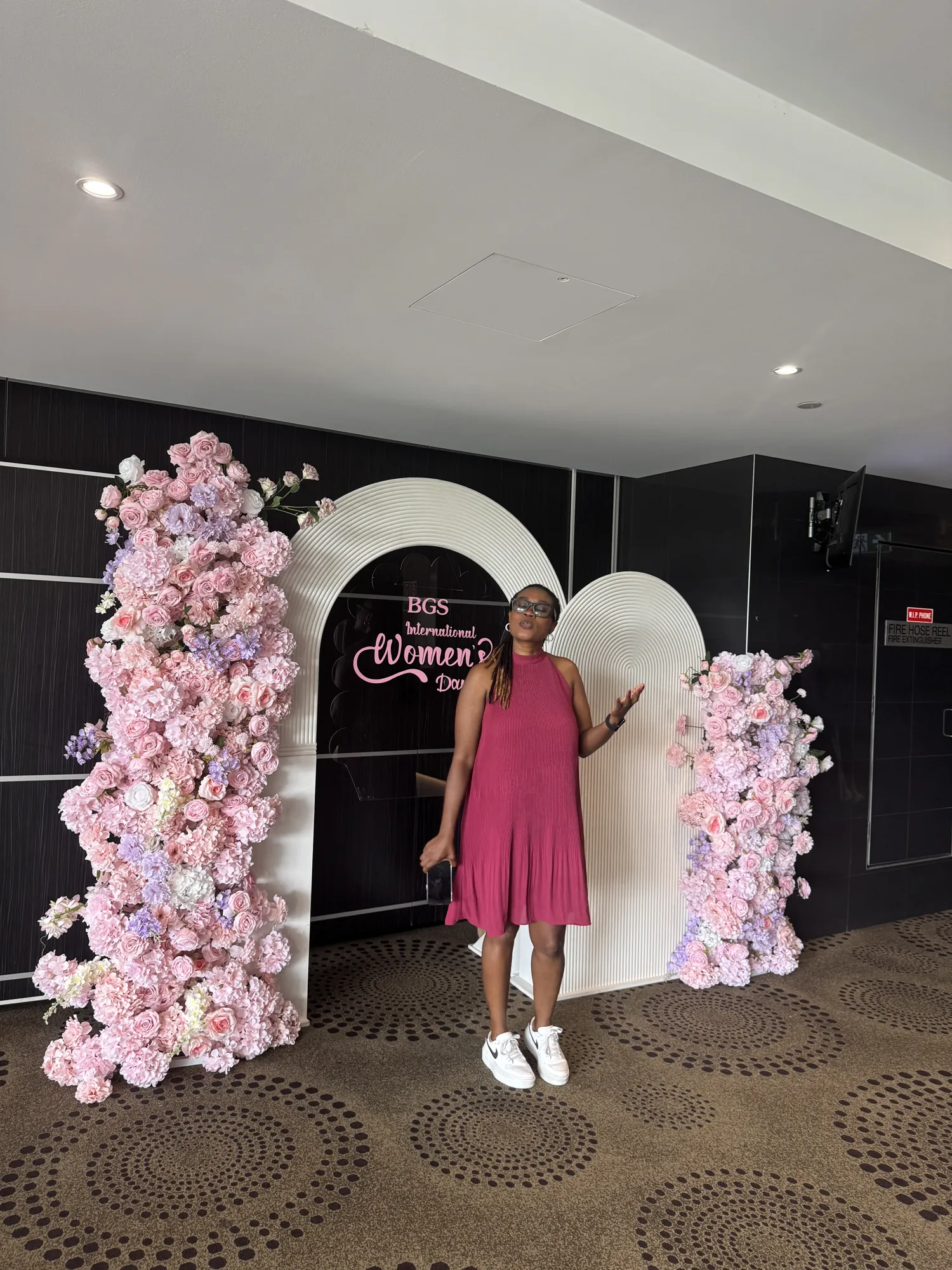A pink floral arch beside a neon sign reading International Women's Day, with a small table of flowers and candles.