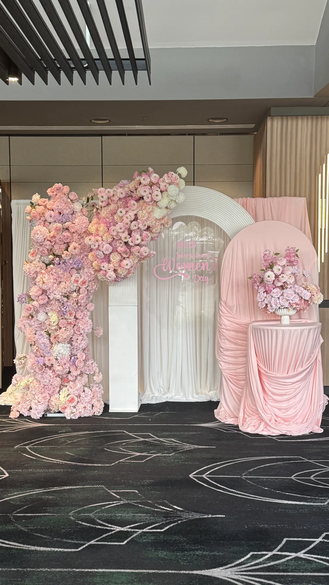 A pink floral arch of roses and greenery, with a glass vase of flowers at its base.