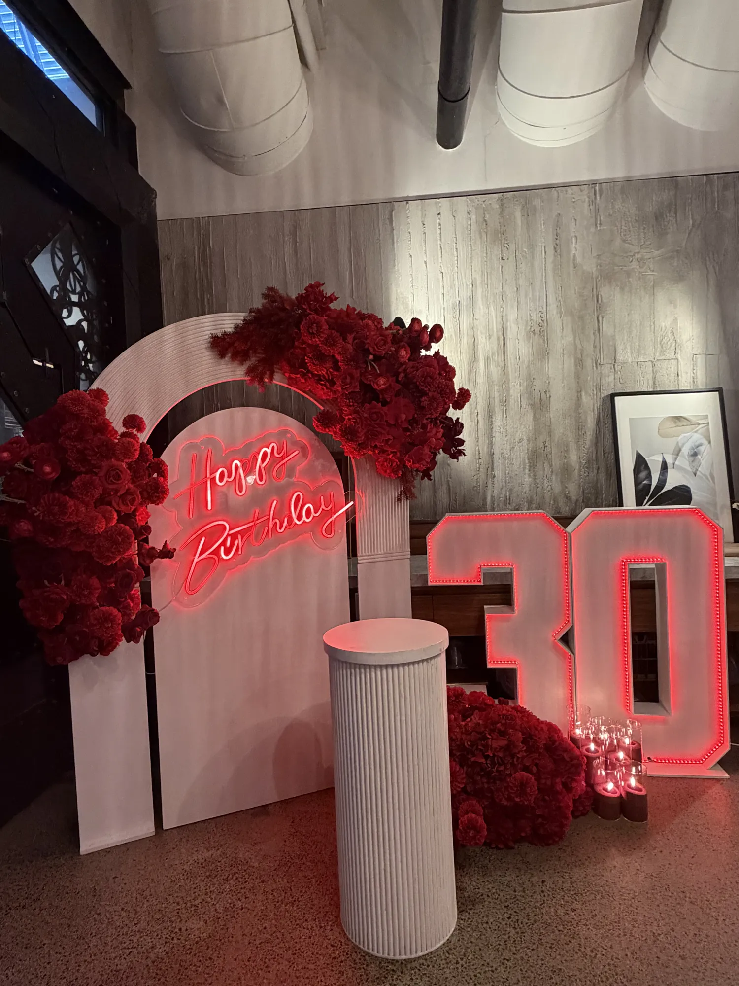 A long table in white linen with a red rose runner, candles, and wicker pendant lights, and a white birthday arch behind.