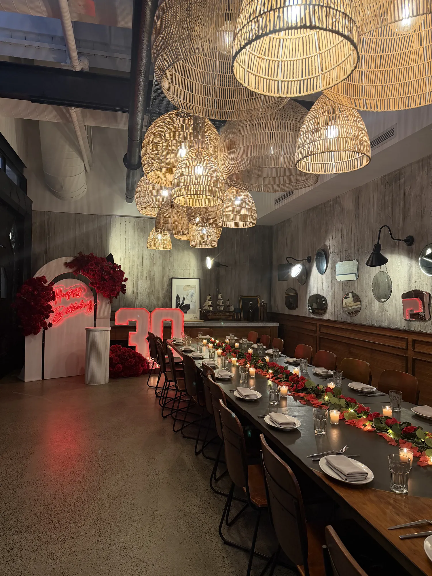 The full length of a table in white linen, with a red rose runner, candles, and wicker pendant lights.