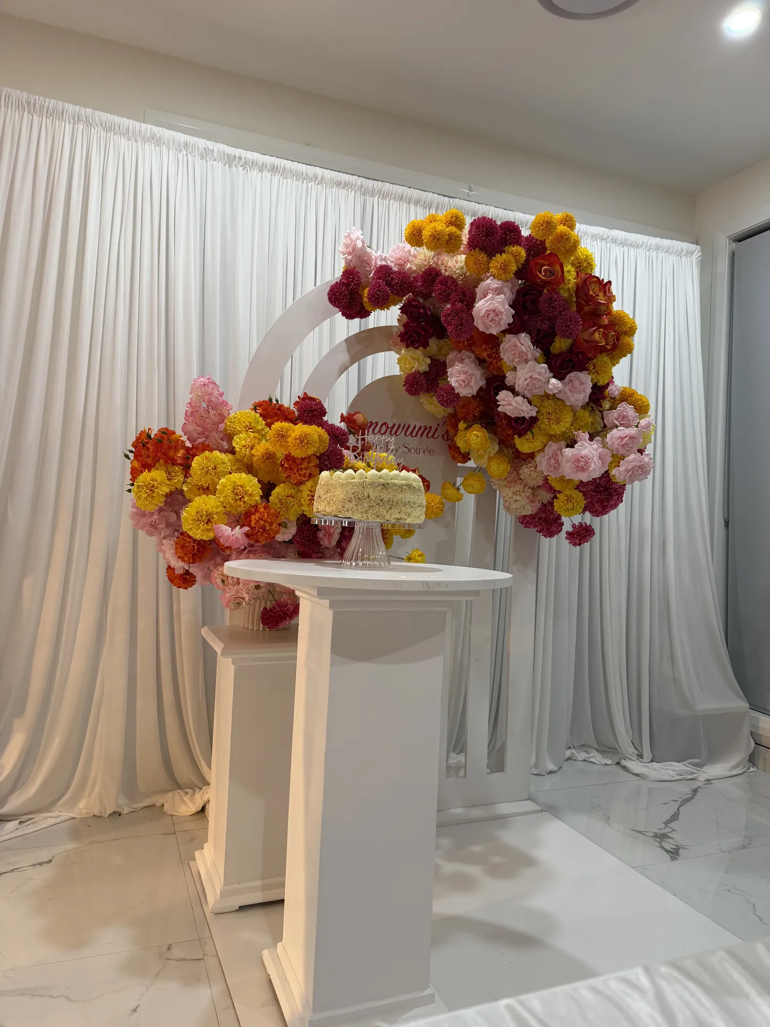 A white floral backdrop with a pink neon sign and a marquee 30, red and white roses, and a three-tier white cake on a glass stand.