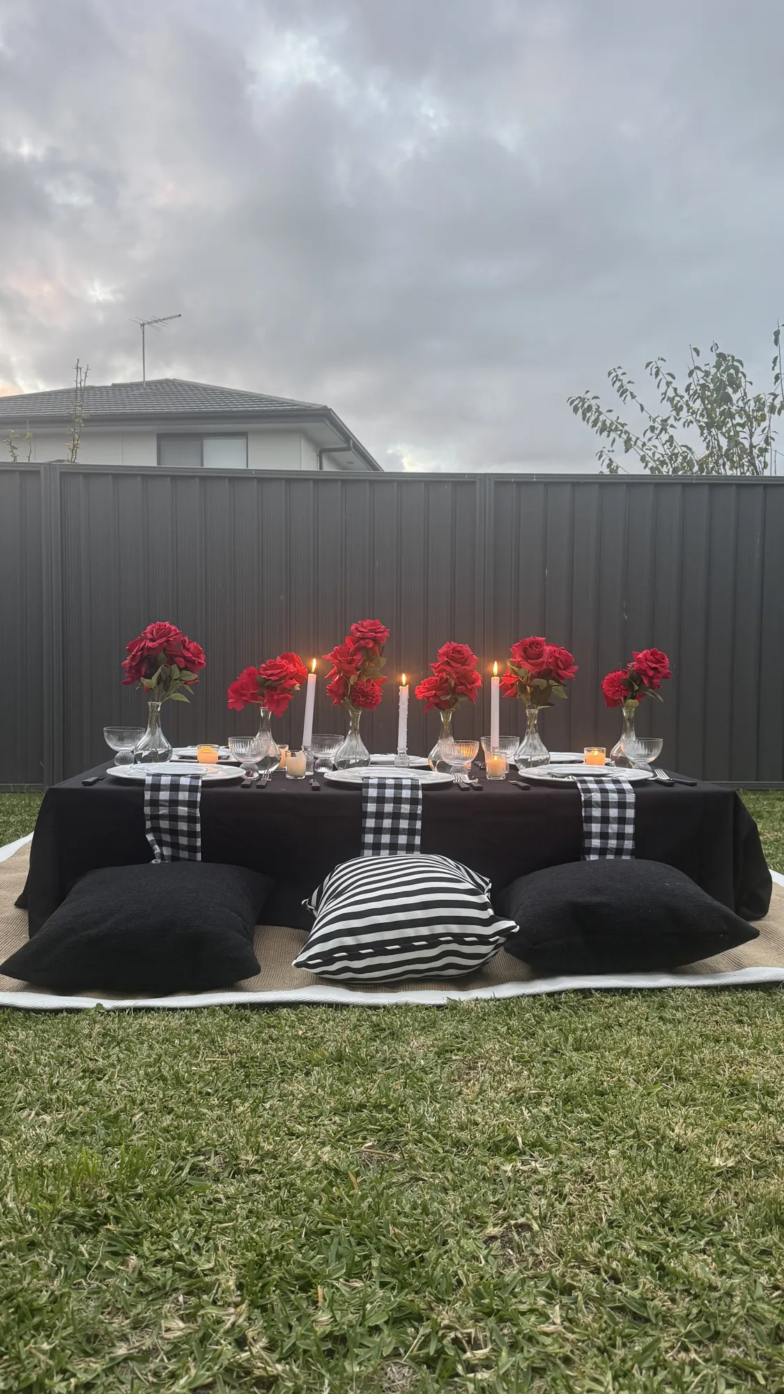 A picnic table in white linen on a lawn, with a white floral centrepiece, a tiered stand, and a white draped backdrop.