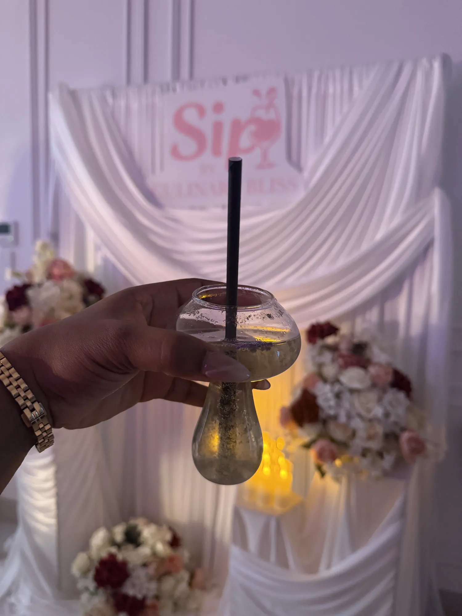 A hand pouring from a clear bottle into a coupe glass, with a neon sign and white flowers behind.
