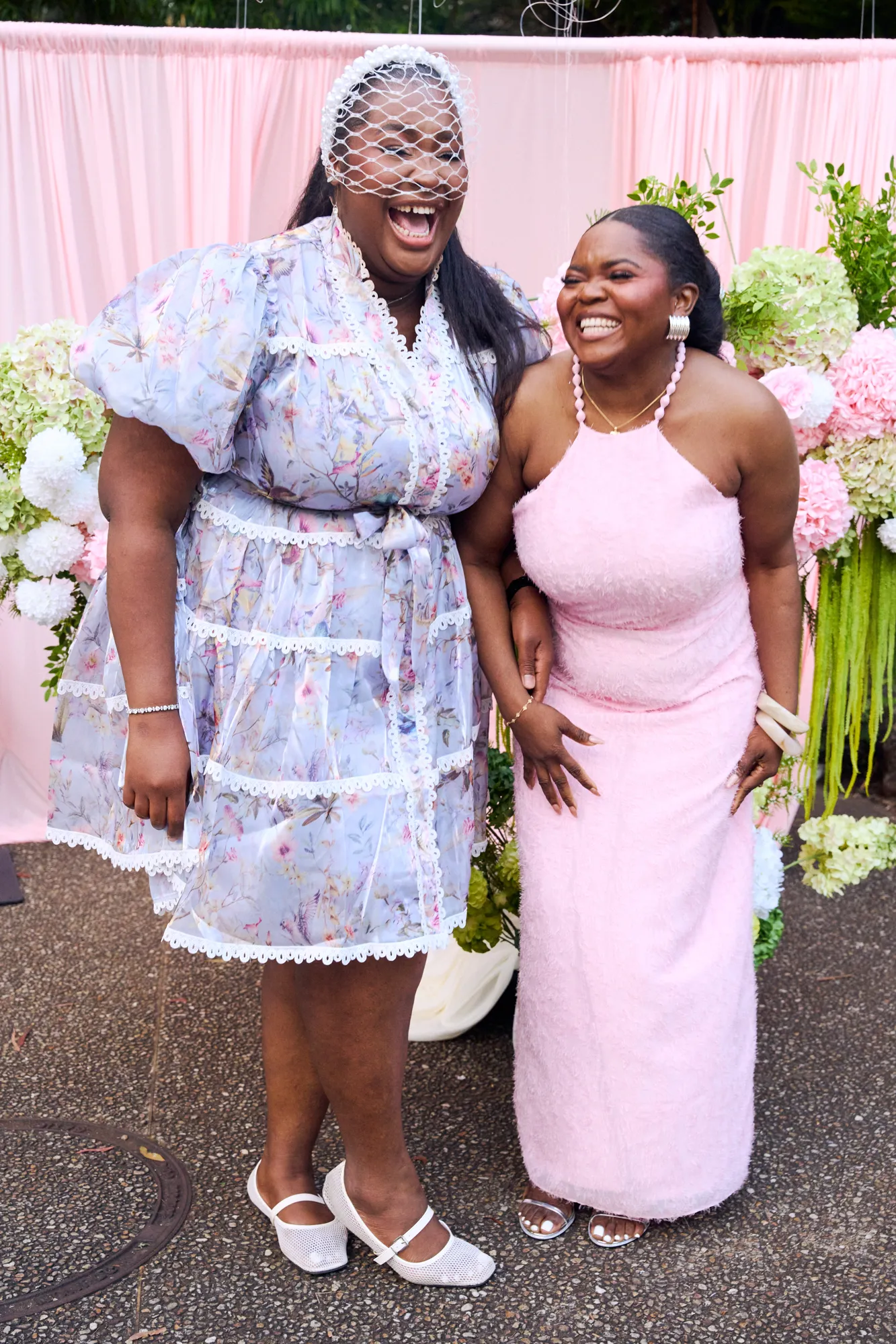 Two guests laughing in front of a white floral backdrop and a pink neon sign.