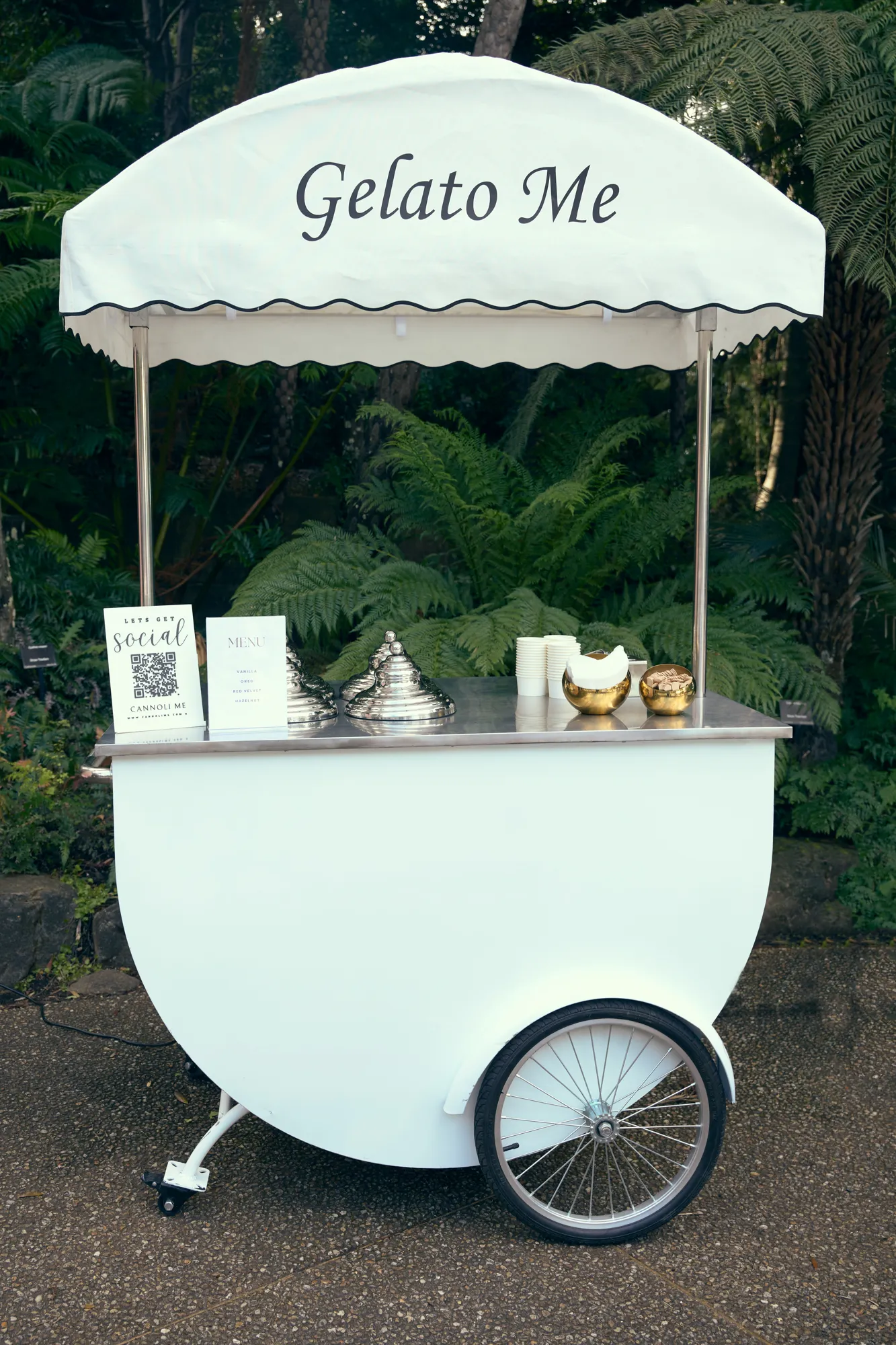 A white Gelato Me cart with a pink and white striped awning, gelato trays, and a vase of pink and white flowers.