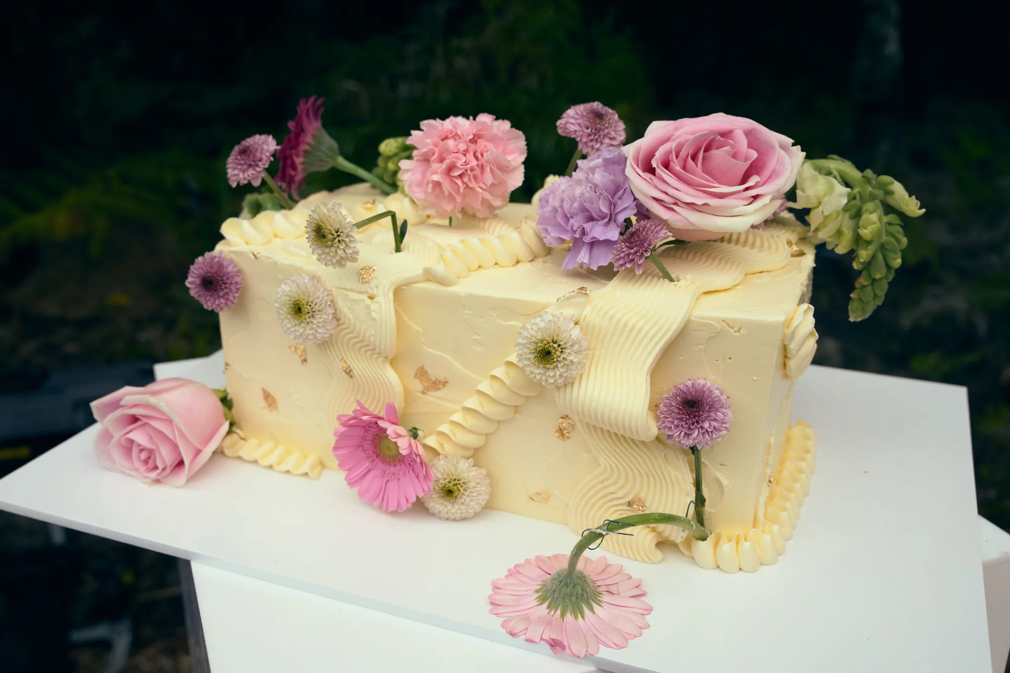 A three-tier white cake with pink and white flowers, gold leaf, and macarons, beside a smaller gold cake.