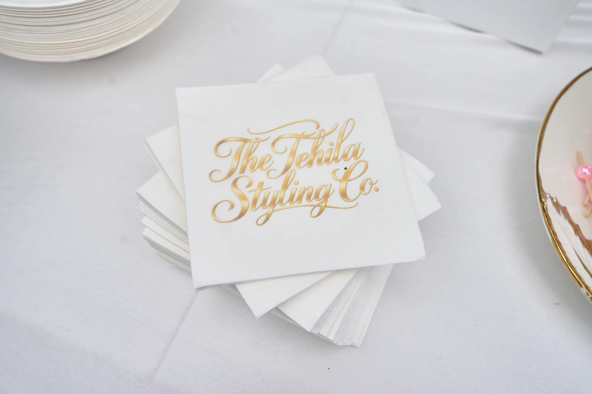 Pink napkins printed with The Tehila Experience, folded on gold charger plates with cutlery and a small white floral bunch.
