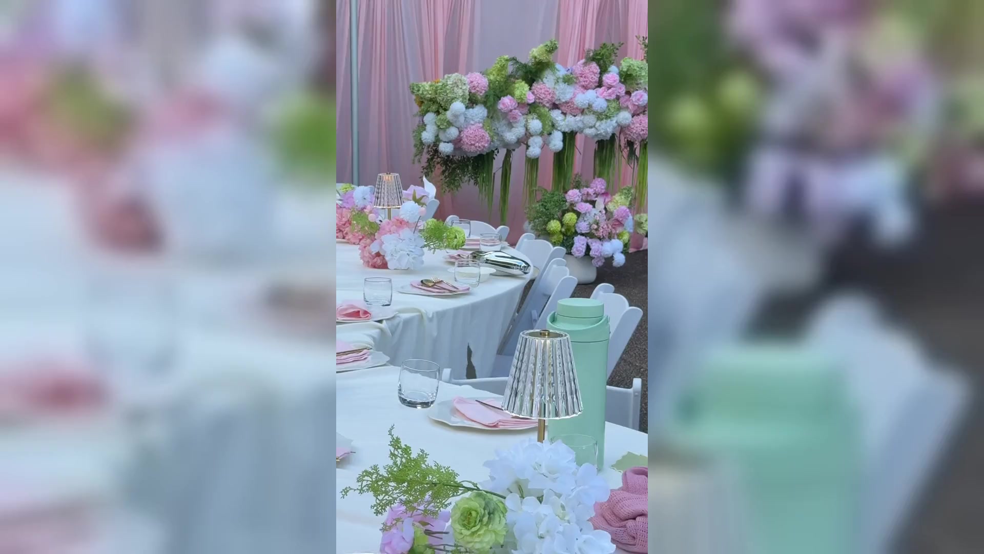 A pink and white floral installation above round tables in white linen, with crystal lamps and pink napkins.