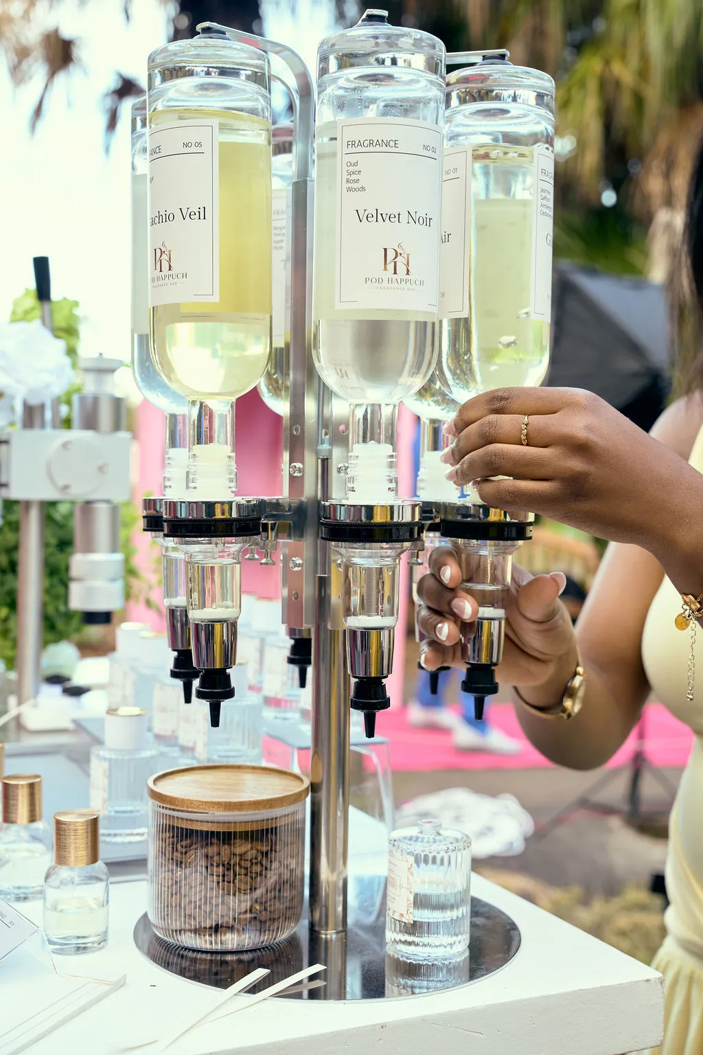 A hand pressing a round gold-top fragrance dispenser, with dark bottles behind and a card reading Petal & Silk.